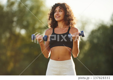 Blue dumbbells are in hands, arms exercise. Young fitness woman is on the field outdoors 137828331