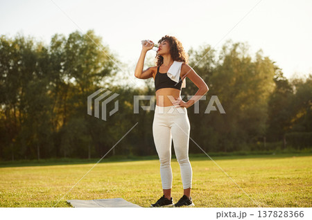 Drinking water from a bottle. Young fitness woman is on the field outdoors Drinking water from a bottle. Young fitness woman is on the field outdoors 137828366