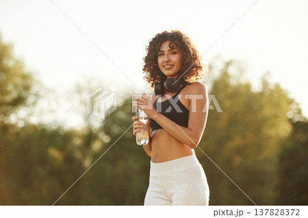Headphones on neck, bottle of water in hand. Young fitness woman is on the field outdoors Headphones on neck, bottle of water in hand. Young fitness woman is on the field outdoors 137828372
