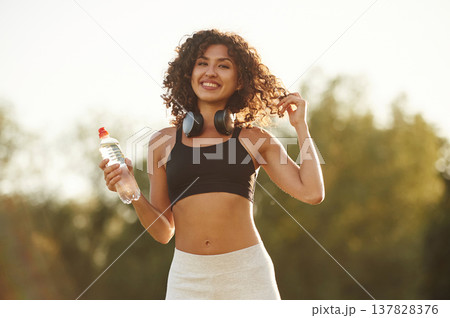Headphones on neck, bottle of water in hand. Young fitness woman is on the field outdoors 137828376