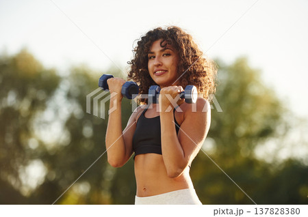 Lifting the dumbbells. Young fitness woman is on the field outdoors 137828380