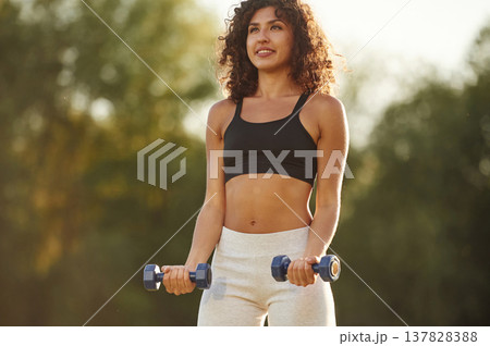 Blue dumbbells are in hands, arms exercise. Young fitness woman is on the field outdoors 137828388