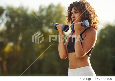 Weight lifting, dumbbells in hands. Young fitness woman is on the field outdoors Weight lifting, dumbbells in hands. Young fitness woman is on the field outdoors 137828400