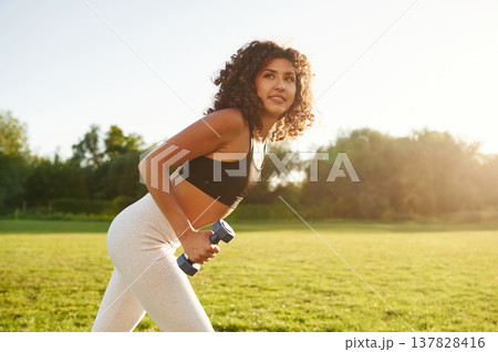 Doing lifting exercises by using dumbbell. Young fitness woman is on the field outdoors 137828416