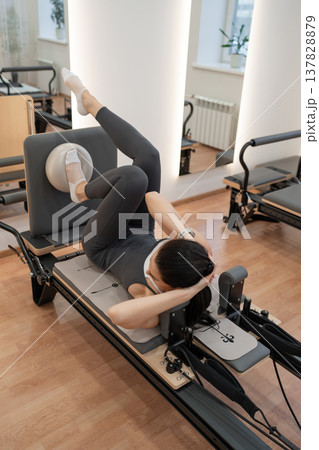 Athletic woman doing reformer crunches with bent knees on mat in white bright studio. Reformer crunches, bent knees, mat, bright studio, athletic. Athletic woman doing reformer crunches with bent knees on mat in white bright studio. Reformer crunches, bent knees, mat, bright studio, athletic. 137828879