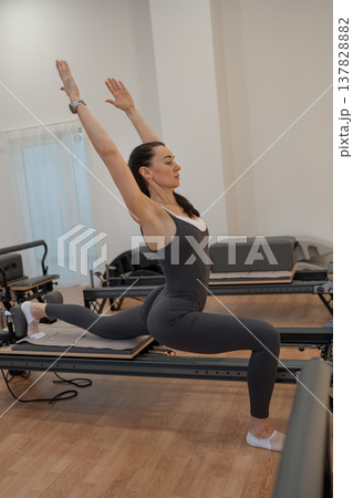 Woman performing pilates with arms high on reformer in white bright studio environment. Arms high, reformer, pilates, bright studio, white. 137828882
