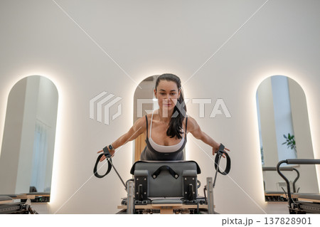 Woman exercising arms on pilates reformer machine in bright studio with mirror walls. Pilates reformer, arm exercise, studio, mirror, bright. 137828901