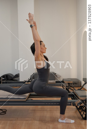Athletic woman on reformer machine, raising arms up in calm yoga pose in bright minimalist studio. Reformer machine, yoga pose, minimalist studio, calm, arms up. Athletic woman on reformer machine, raising arms up in calm yoga pose in bright minimalist studio. Reformer machine, yoga pose, minimalist studio, calm, arms up. 137828903