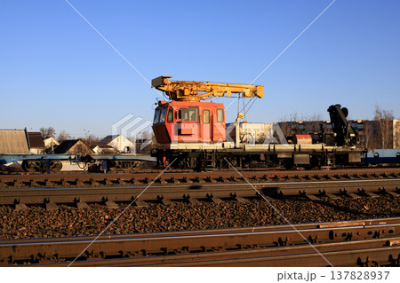 Heavy railway track-laying crane stands on the tracks near a train station under a clear blue sky. Heavy railway track-laying crane stands on the tracks near a train station under a clear blue sky. 137828937