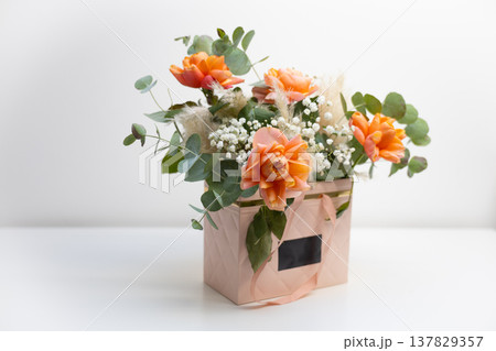 Close view of orange roses and delicate filler flowers arranged with eucalyptus in a pastel floral gift box. Romantic decoration, botanical elegance, festive surprise, and stylish flower arrangement 137829357