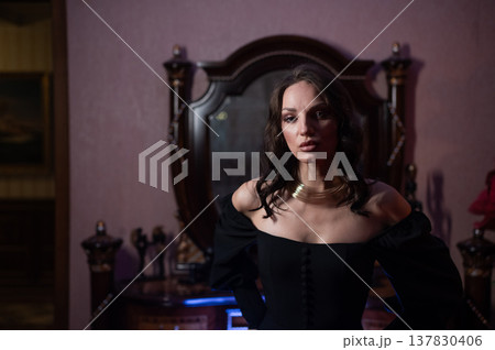 A beautiful brunette in a black evening dress poses in front of a retro-style mirror. A beautiful brunette in a black evening dress poses in front of a retro-style mirror. 137830406