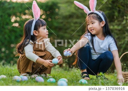 two child girl wearing bunny ears playing together and hunting for Easter eggs into basket on green grass in garden two child girl wearing bunny ears playing together and hunting for Easter eggs into basket on green grass in garden 137836916