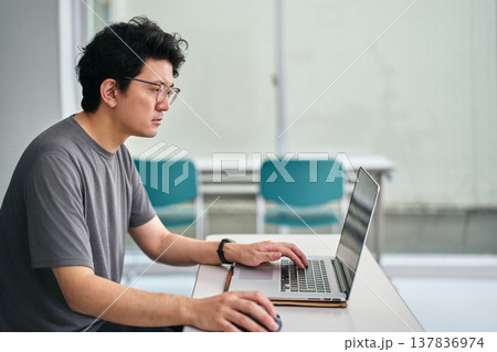 室内でTシャツを着てパソコンを使い仕事をする眼鏡をかけた男性 室内でTシャツを着てパソコンを使い仕事をする眼鏡をかけた男性 137836974