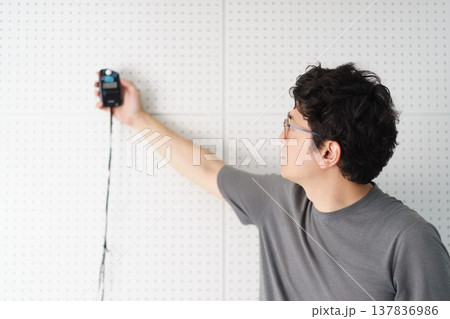 吸音ボードの貼られた室内で光の強さを測定する露出計を使う眼鏡をかけた1人の男性 137836986