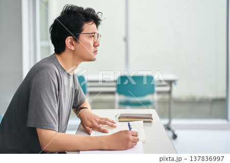 講義を受ける教室で教科書を手元にノートでメモを取りながら真剣に授業を聞く男性の学生 137836997