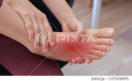 Woman sitting on floor and massaging her painful ankle with red highlight. Ankle sprain, joint inflammation, tendonitis and injury concept for health care, physical therapy and orthopedic treatment. 137838845