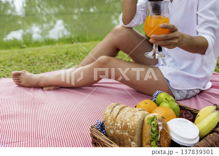 Close up Women picnic in green park hands holding glass of orange juice fresh fruit cool summer. Women drinking sit blanket with Picnic basket bread coffee cup in wood wicker healthy box red Blanket Close up Women picnic in green park hands holding glass of orange juice fresh fruit cool summer. Women drinking sit blanket with Picnic basket bread coffee cup in wood wicker healthy box red Blanket 137839301