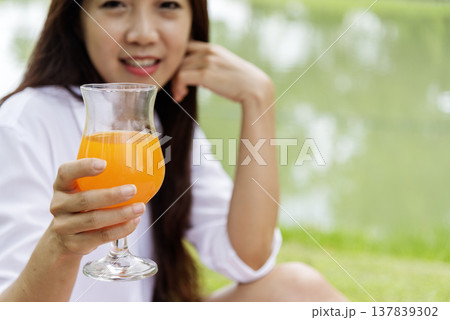 Women hands drink orange juice fresh fruit cool drinking in green park garden. Asian women hands holding healthy drinking glass. Beautiful female pouring orange juice from cocktail bottle outdoors Women hands drink orange juice fresh fruit cool drinking in green park garden. Asian women hands holding healthy drinking glass. Beautiful female pouring orange juice from cocktail bottle outdoors 137839302