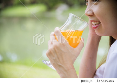 Close up Women picnic in green park hands holding glass of orange juice fresh fruit cool summer. Women drinking sit blanket with Picnic basket bread coffee cup in wood wicker healthy box red Blanket Close up Women picnic in green park hands holding glass of orange juice fresh fruit cool summer. Women drinking sit blanket with Picnic basket bread coffee cup in wood wicker healthy box red Blanket 137839303