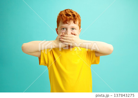 Little Red Headed Boy Covers Mouth With Hands, Looking to Camera, doesn't Want Say Anything, Wearing in Yellow T-shirt Standing Over Blue Isolated Little Red Headed Boy Covers Mouth With Hands, Looking to Camera, doesn't Want Say Anything, Wearing in Yellow T-shirt Standing Over Blue Isolated 137843275