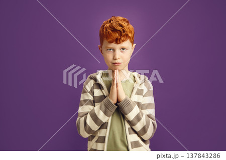 Celtic appearance little boy with red head has curly hair keeping hands in palm, symbolizing gesture of supplication, asking for something very much, as if saying please standing on violet isolated Celtic appearance little boy with red head has curly hair keeping hands in palm, symbolizing gesture of supplication, asking for something very much, as if saying please standing on violet isolated 137843286