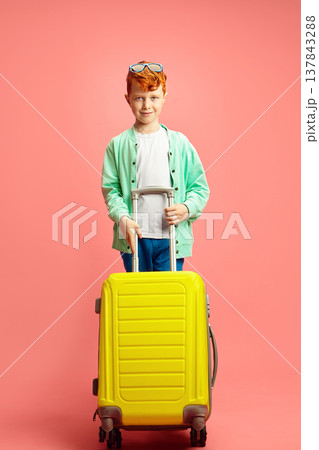 Handsome red haired little boy holding yellow suitcase, wearing in sunglasses on head, dressed in colorful casual clothes standing on pink isolated background. 137843288