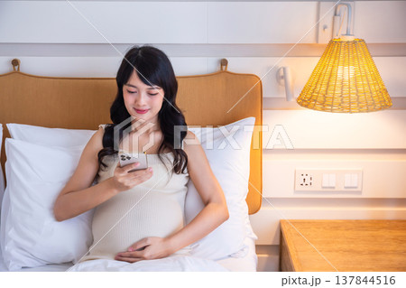 Pregnant woman uses smartphone to search for baby news on bed in bedroom with warm lamp and hands gently stroking belly. 137844516