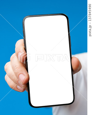 Hand holding a smartphone with a blank screen against a blue background in a simple setting 137845490