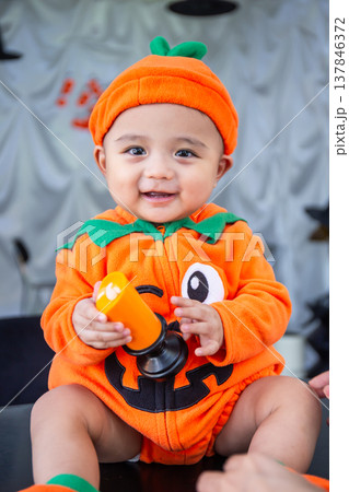 A little boy wearing an orange pumpkin costume and hat sits smiling happily on a table holding candles to celebrate Halloween. A little boy wearing an orange pumpkin costume and hat sits smiling happily on a table holding candles to celebrate Halloween. 137846372