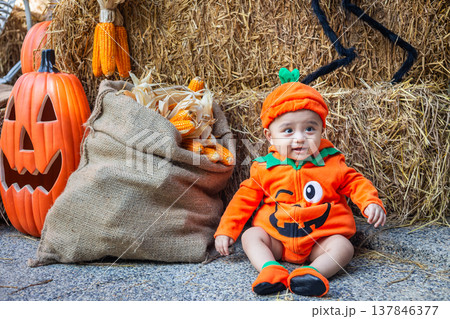 A little boy wearing a pumpkin costume and hat sits on a woven cloth to celebrate Halloween with a background of pumpkins, ghosts and skeletons. A little boy wearing a pumpkin costume and hat sits on a woven cloth to celebrate Halloween with a background of pumpkins, ghosts and skeletons. 137846377