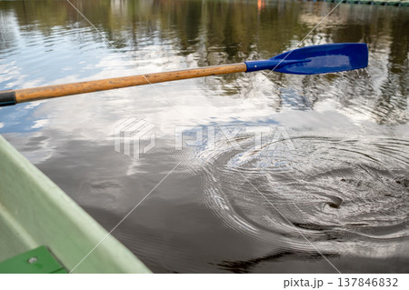 A blue paddle stirs the still water, creating ripples under a cloudy sky as the afternoon light reflects off the calm lake surface surrounded by nature. 137846832