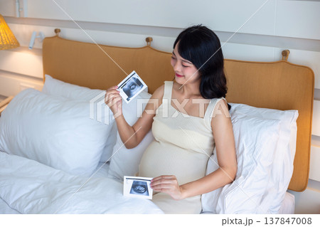 Pregnant woman sitting on bed holding ultrasound image of baby with happy smile 137847008