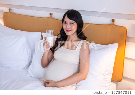 Pregnant woman lying on bed in bedroom drinking milk for health of both mother and baby with love and warmth. Pregnant woman lying on bed in bedroom drinking milk for health of both mother and baby with love and warmth. 137847011