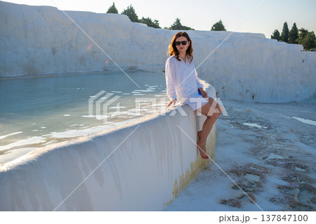 A woman enjoys a calm moment by the thermal pools in Pamukkale, surrounded by stunning white travertine terraces, under the warm evening sun. A woman enjoys a calm moment by the thermal pools in Pamukkale, surrounded by stunning white travertine terraces, under the warm evening sun. 137847100