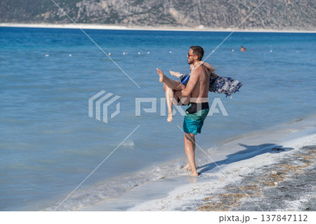 Man carries woman playfully along the shore, embracing warmth and joy by the tranquil ocean under a bright blue sky on a sunny summer day. Man carries woman playfully along the shore, embracing warmth and joy by the tranquil ocean under a bright blue sky on a sunny summer day. 137847112