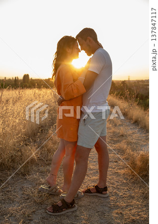 Two people share a loving moment while the sun sets behind them, creating a beautiful silhouette against a tranquil landscape of golden grass. Two people share a loving moment while the sun sets behind them, creating a beautiful silhouette against a tranquil landscape of golden grass. 137847117