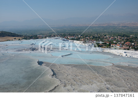 Pamukkale showcases its famous white travertine terraces and hot springs under a bright sky, with a picturesque village visible in the background. 137847161