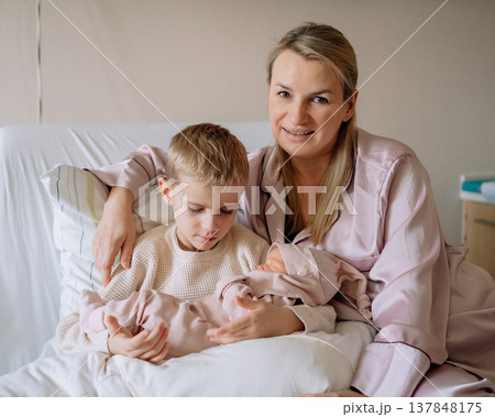 Mother holding newborn baby while sitting on a hospital bed with her older son beside her in a hospital room after childbirth. Tender family moment during the first days of baby life. Mother holding newborn baby while sitting on a hospital bed with her older son beside her in a hospital room after childbirth. Tender family moment during the first days of baby life. 137848175