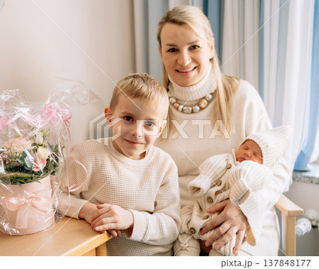 Mother holding newborn baby while older child sits next to her in hospital room after birth. Warm emotional moment of motherhood and family bonding. 137848177