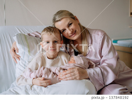 Young mother caring for newborn baby on hospital bed while older child sits nearby in hospital room. Concept of motherhood, family bonding and first days after birth. 137848192