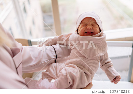 Newborn baby crying while being held by mother near window light. Natural moment of infant emotions during first days after birth. Newborn baby crying while being held by mother near window light. Natural moment of infant emotions during first days after birth. 137848238