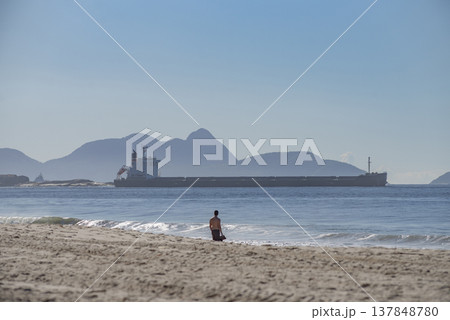 Lone Man on Sandy Beach Watching Large Cargo Ship with Mountain Background and Copy Space 137848780
