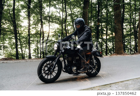 Bunch of trees behind. Man in leather clothes on motorcycle is on the road Bunch of trees behind. Man in leather clothes on motorcycle is on the road 137850462
