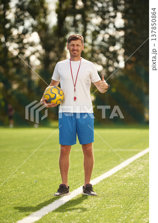 Happy smiling soccer trainer is standing on the field with ball in hand, showing thumb up Happy smiling soccer trainer is standing on the field with ball in hand, showing thumb up 137850864