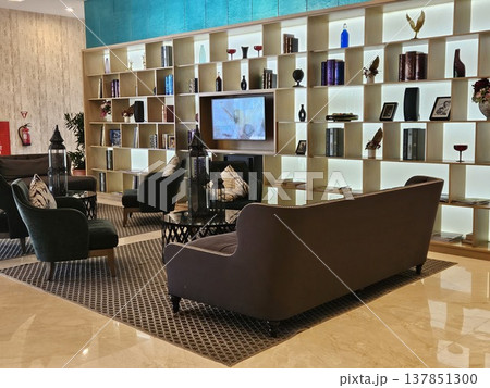 A sophisticated hotel lounge featuring a large illuminated bookshelf wall, plush dark velvet sofas, and elegant decor items, creating a warm and luxurious atmosphere for guests in a modern interior. A sophisticated hotel lounge featuring a large illuminated bookshelf wall, plush dark velvet sofas, and elegant decor items, creating a warm and luxurious atmosphere for guests in a modern interior. 137851300