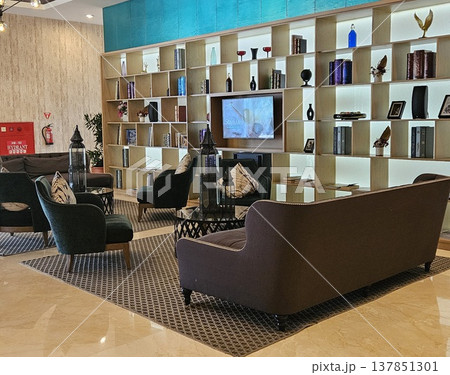 A sophisticated hotel lounge featuring a large illuminated bookshelf wall, plush dark velvet sofas, and elegant decor items, creating a warm and luxurious atmosphere for guests in a modern interior. A sophisticated hotel lounge featuring a large illuminated bookshelf wall, plush dark velvet sofas, and elegant decor items, creating a warm and luxurious atmosphere for guests in a modern interior. 137851301