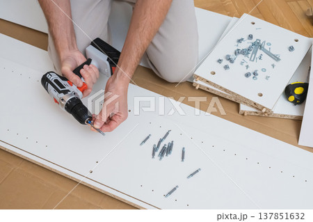 Male hands use a screwdriver to fasten hardware into a white furniture panel with assembly parts and instructions laid out on a wooden floor Male hands use a screwdriver to fasten hardware into a white furniture panel with assembly parts and instructions laid out on a wooden floor 137851632