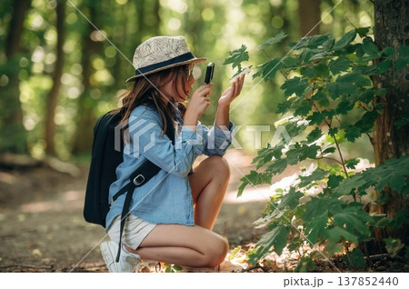 Discovering plants under magnifying glass. Young girl hiker is in the forest Discovering plants under magnifying glass. Young girl hiker is in the forest 137852440
