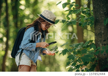 Discovering plants under magnifying glass. Young girl hiker is in the forest 137852444