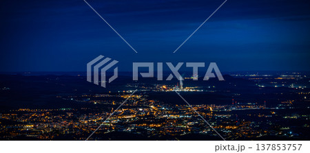Aerial view of a city illuminated at night with glowing lights, distant mountains, and a power plant emitting steam in the background under a twilight sky Aerial view of a city illuminated at night with glowing lights, distant mountains, and a power plant emitting steam in the background under a twilight sky 137853757